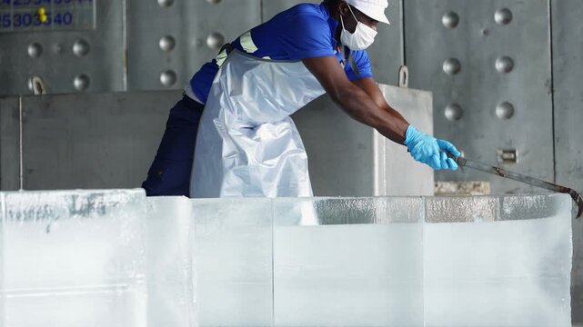 Masked African worker in a white cap bends over a row of clear ice blocks and drags one with a long steel hook, his silver apron and rubber boots visible as he walks