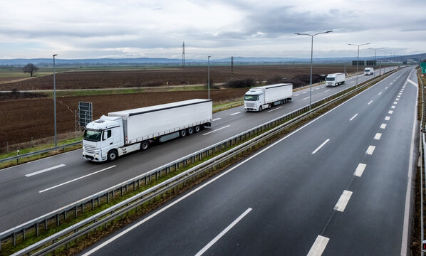 An elevated view of a line of white semi-trailer trucks driving along a multi-lane highway through a rural landscape under an overcast sky.