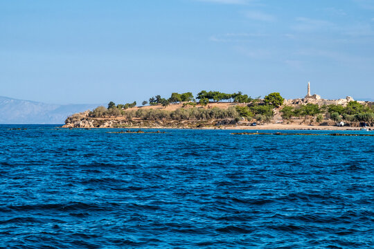 Panoramic view across deep blue sea waters toward the coastal Kolona Hill on Aegina Island, featuring pine trees and the lone remaining column of the ancient Temple of Apollo.
