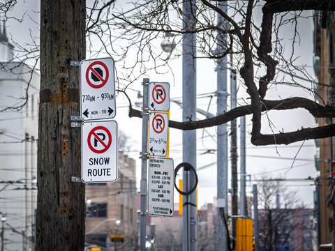 American Parking regulation signs in Toronto, Canada, showing no parking symbols, snow route information and timed curbside restrictions on a winter urban street with utility poles and bare trees.