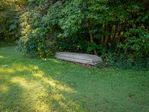 An overturned metal boat resting on grass near dense green trees in a sunny park setting