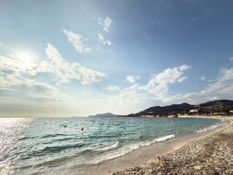 Panorama of Plage de l'Almanarre in Hyeres, France, showing a Mediterranean beach, calm water, sandy shoreline and green hills with scattered homes along the coast of the French Riviera.