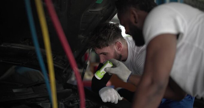 Two mechanics inspecting car air conditioning system with manifold gauge and flashlight