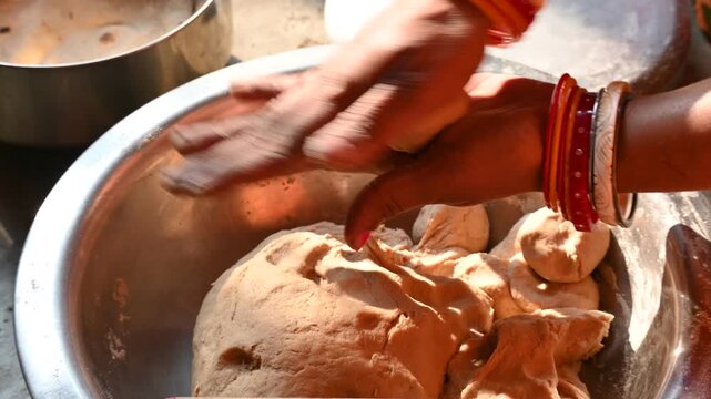 Woman shaping whole wheat dough for roti in a metal bowl. A pair of women hands wearing colorful bangles kneads and shapes soft brown dough inside a large metallic bowl, with additional kitchen.