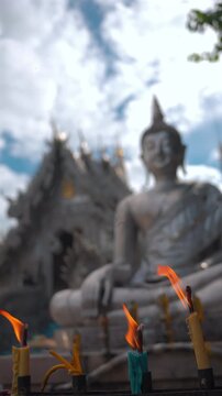 burning colorful candles in front of blurred silver buddha statue, lit fire offerings near large metallic religious sculpture outside, vertical video 4k