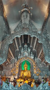 golden buddha statue inside ornate Silver Temple interior, sunlight illuminating intricate carved metal architecture of traditional Thai shrine Wat Sri Suphan, 4k vertical video