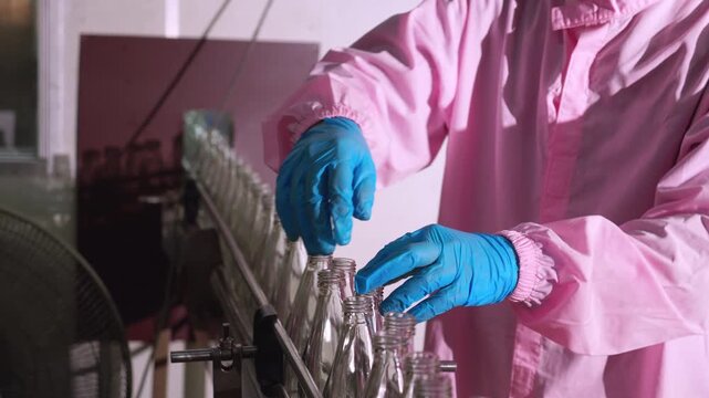 Worker hands wearing gloves placing empty glass bottles on industrial automated conveyor belt line inside modern  beverage factory production plant facility, manufacturing process in industry.
