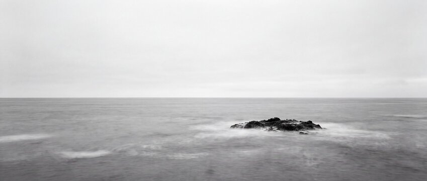 Lonely rock formation standing alone in vast calm ocean water under overcast moody sky