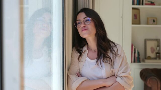 Woman relaxes at home by a bright window, looking outside and toward the camera with a gentle smile. The young woman pauses thoughtfully beside a bookshelf in a sunlit, comfortable living room.