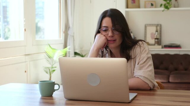 Woman works on a laptop from home, gesturing and pausing thoughtfully while looking at the screen. Young freelancer in glasses sits at a wooden table in a bright living room with natural window light.