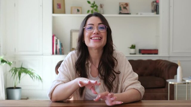 Woman talks directly to the camera, smiles and gestures during a video call at home. The young woman wears glasses at a wooden table in a bright home office, speaking with a friendly, warm mood.