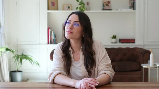Woman works from home at a wooden desk, looking up and resting her chin on her hand thoughtfully. Young freelancer pauses in a bright living room home office during remote work, study, or planning.