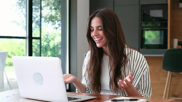 Woman talks and gestures during a laptop video call at home, smiling warmly toward the screen. She sits at a wooden table in daylight beside a notebook and pen, with kitchen cabinets behind her.