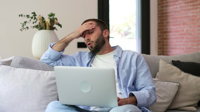 Stressed young man works on a laptop while seated on a sofa at home, then sighs and rubs his forehead in frustration. Tired remote worker faces workload, burnout, fatigue and mental health pressure.