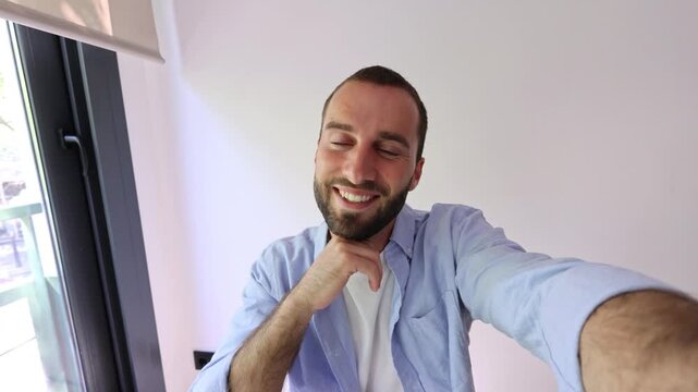 Man talks directly to the camera, pointing, gesturing and waving during a video call from home. Selfie content shows the bearded young man in a bright room with window light, white walls and blinds.