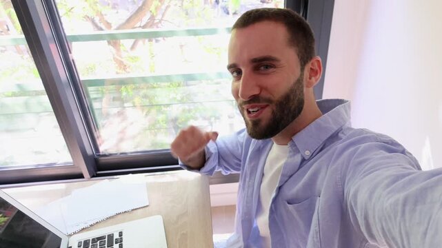 Young man talks directly to the camera while typing on a laptop at a desk near a large window at home. He records a selfie video, smiles broadly, and raises his fist in a cheerful, positive.