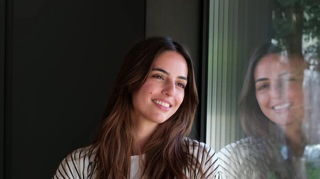 Woman smiles and gazes out the window at home, enjoying a quiet moment in soft daylight. Her reflection appears on the glass pane as she looks upward, thoughtful and hopeful in a bright interior.