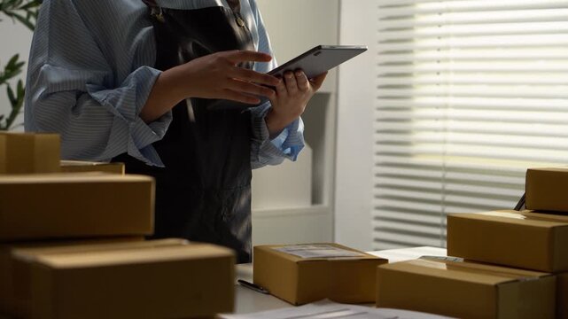 A seller uses a tablet beside stacked parcels in a packing area. Cardboard boxes and digital tools illustrate ecommerce order management, small business shipping, and preparation for parcel delivery.