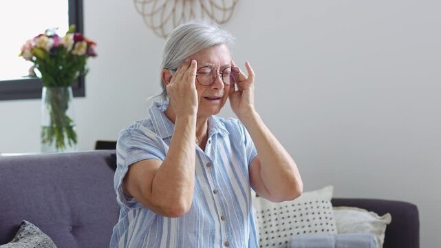Senior woman massages her temples with eyes closed as severe headache pain continues while sitting at home. Static medium shot shows migraine, illness, stress, senior health and healthcare.