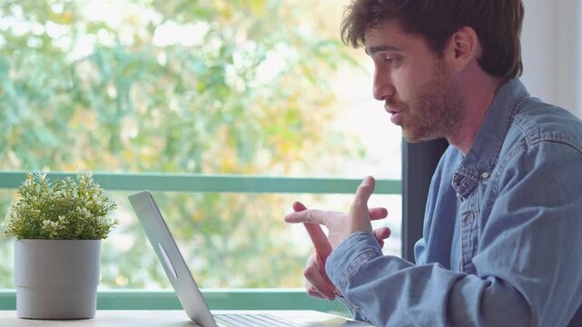 Man talks and gestures to a laptop during a remote work video call at home. Young freelancer sits at a desk, sharing ideas in an online meeting with daylight and trees visible through the window.