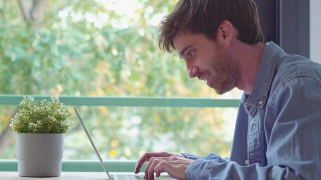 Smiling man writes notes in a notebook at a table by the window before setting down his pen and typing on a laptop. Static side-profile shot shows remote work, study, and freelance tasks at home.