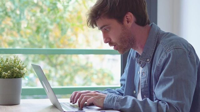Man works from home by writing notes on paper, then typing on a laptop at a wooden desk. The focused young freelancer checks his screen beside a potted plant and window in a bright home office.