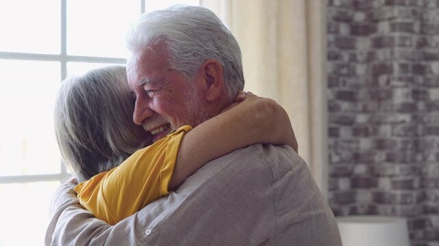 Senior couple hug and smile together at home, sharing love, romance and affection. Elderly man pulls his partner close by a bright window in a tender embrace, expressing marriage and companionship.