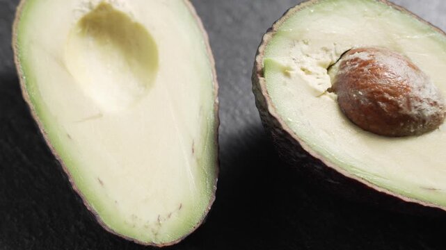 High angle close-up of two avocado halves on a dark slate surface, one with the brown pit, as the camera moves slowly across them.