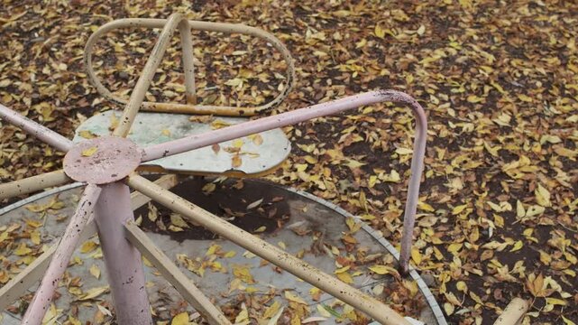 Medium shot of a weathered vintage metal carousel spinning counterclockwise on a ground covered with autumn leaves in a park in slow motion