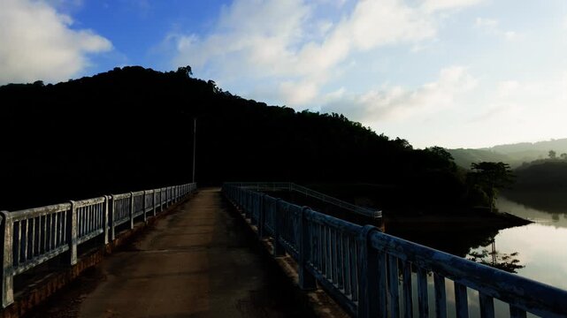 At Da Lay Irrigation Reservoir, the sun rises over stunning hills. Water reflects the sky as people walk along the bridge. Nature unfolds in Lam Dong, Vietnam.