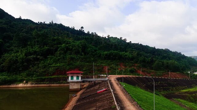People visit Da Lay Irrigation Reservoir in Lam Dong, Vietnam. They walk by the water and take pictures of the area. The sky is cloudy and the green hills surround the reservoir.