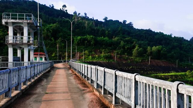 At Da Lay Irrigation Reservoir in Lam Dong, Vietnam, a wide path runs alongside the water. Tall trees dot the hillside, adding to the view. The bright sky enhances the scenery.
