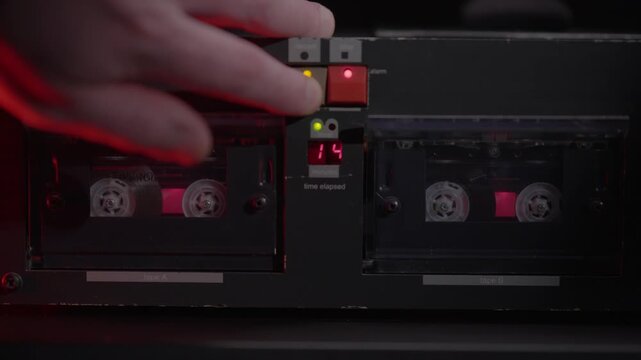 Hand loads a cassette tape into a dual deck player under red light. Gritty retro audio recording scene, ideal for police interrogation, wiretap evidence, crime mystery, and forensic thrillers.