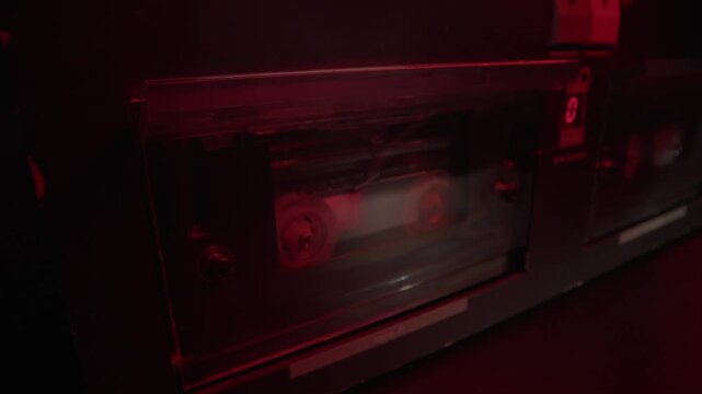 Hand loads a cassette tape into a dual deck player under red light. Gritty retro audio recording scene, ideal for police interrogation, wiretap evidence, crime mystery, and forensic thrillers.