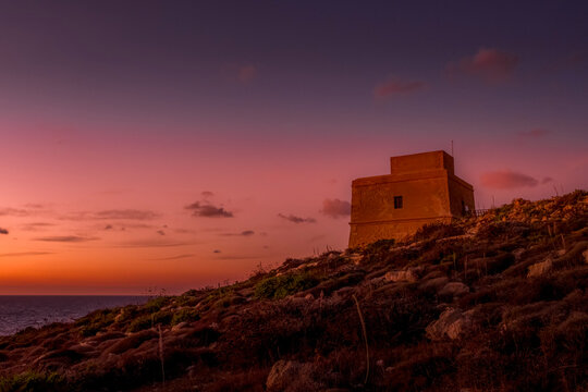 Historic Stone Watchtower on Coastal Hillside Overlooking Sea under Pastel Pink, Purple and Orange Sunset Sky, Dwejra