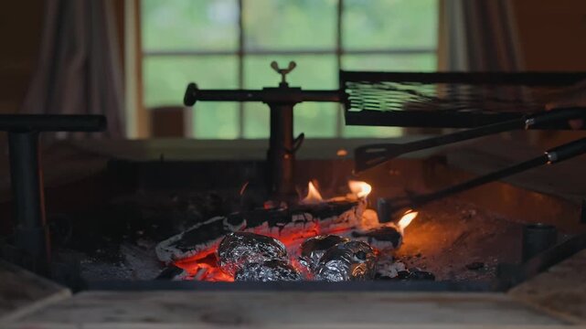 Close-up of aluminium foil-wrapped potatoes roasting among glowing embers and flames in a traditional fireplace as metal fire tongs turn them in a warm, rustic cooking scene.