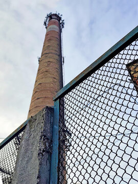 Industrial chimney viewed from below behind a metal mesh fence, with a brick building and cloudy sky in the background. Urban decay and industrial architecture concept.
