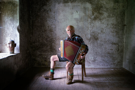 Senior musician playing traditional accordion sitting on chair in abandoned rustic room