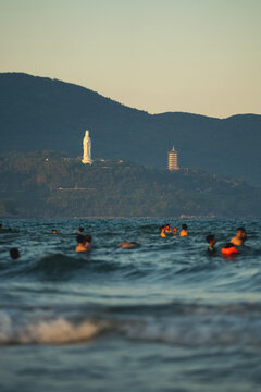 People swim in Danang sea, Lady Buddha statue and Linh Ung Pagoda above waves and tourists in Da Nang, Coastal view of Son Tra Peninsula with Buddhist landmark, mountains and ocean shoreline