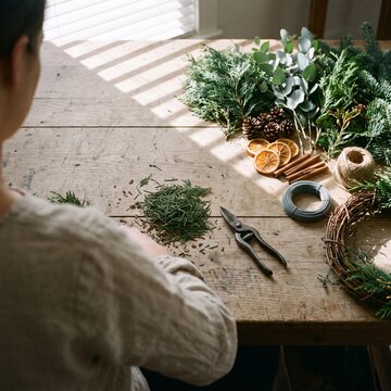 Over the shoulder view of a woman crafting a traditional Christmas wreath on a rustic wooden table with pine branches, dried oranges, and cinnamon sticks