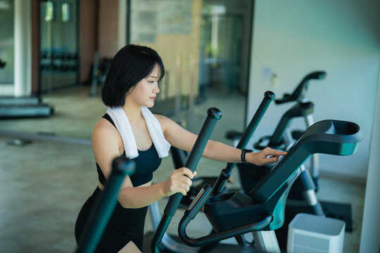 Young woman exercising on elliptical machine in modern gym wearing sportswear with towel around neck and focused expression