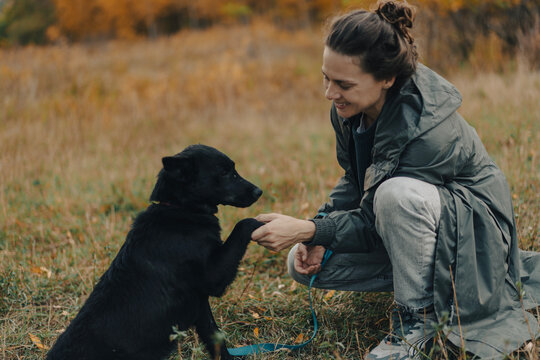 Smiling young woman in raincoat holding her black dog paw during autumn walk