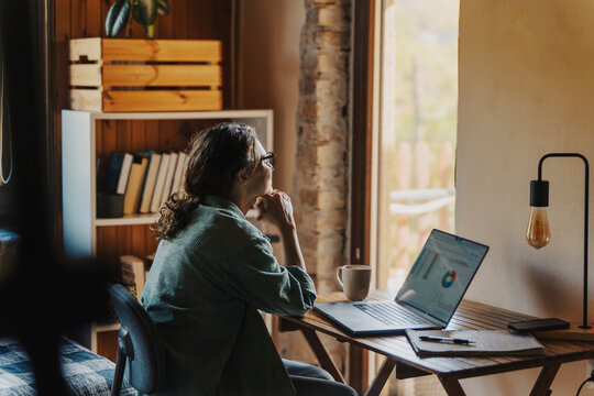 A back view over the shoulder shot of a smiling young woman around 30 years old sitting at her home office desk with a laptop and looking out the window
