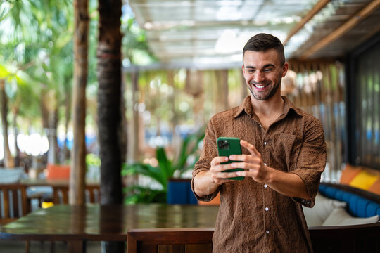 Happy male digital nomad checking smartphone while enjoying vacation at tropical resort