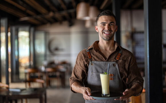 Young smiling handsome man waiter serving fruit juice, smoothie while working in a cafe.