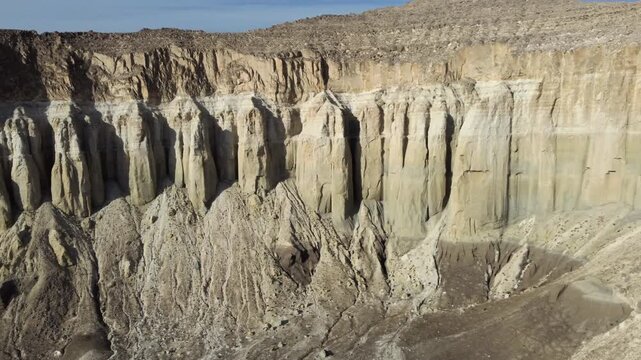 Drone glides laterally past a sunlit escarpment of vertical limestone columns and grey talus cones at the base of the Airakty cliffs