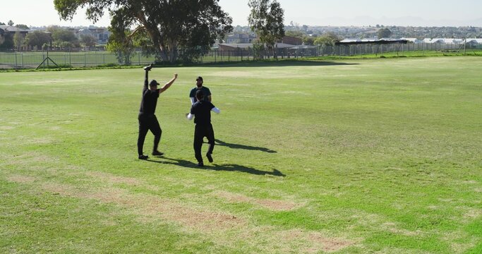 Three male recreational cricketers celebrating on sports field in sportswear holding bat and gloves
