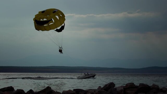 Motorboat towing yellow smiley face parasail with two people gliding through air above sea water.