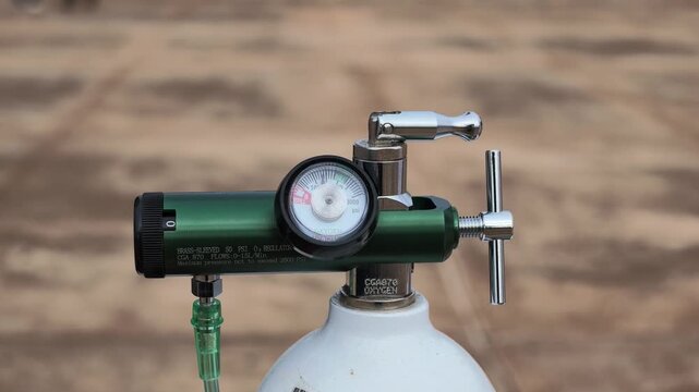 Green medical oxygen regulator with pressure gauge, flow knob and tubing on a white portable oxygen cylinder against a blurred wooden background, slow orbit