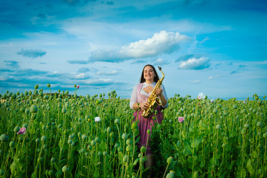 Smiling young woman in traditional dirndl holding golden saxophone in green poppy seed field